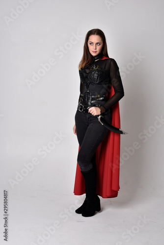 full length portrait of a pretty brunette woman wearing black leather fantasy costume with long red superhero cape. standing pose on a studio background.
