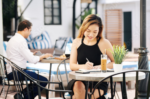 Beautiful young Vietnamese business lady drinking iced coffee and writing down plans in diary