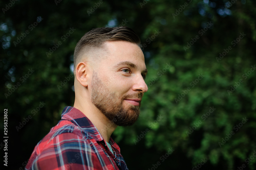 Brutal bearded young man in red shirt is standing outdoors on green trees background. Hipster style concept. Brutal bearded young man in red shirt is standing outdoors on green trees background. Hipster style concept.
