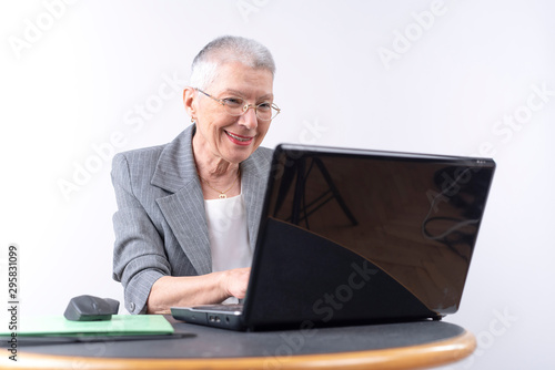 Senior old business lady, sitting at her office table and typing on her laptop, looking cheerful