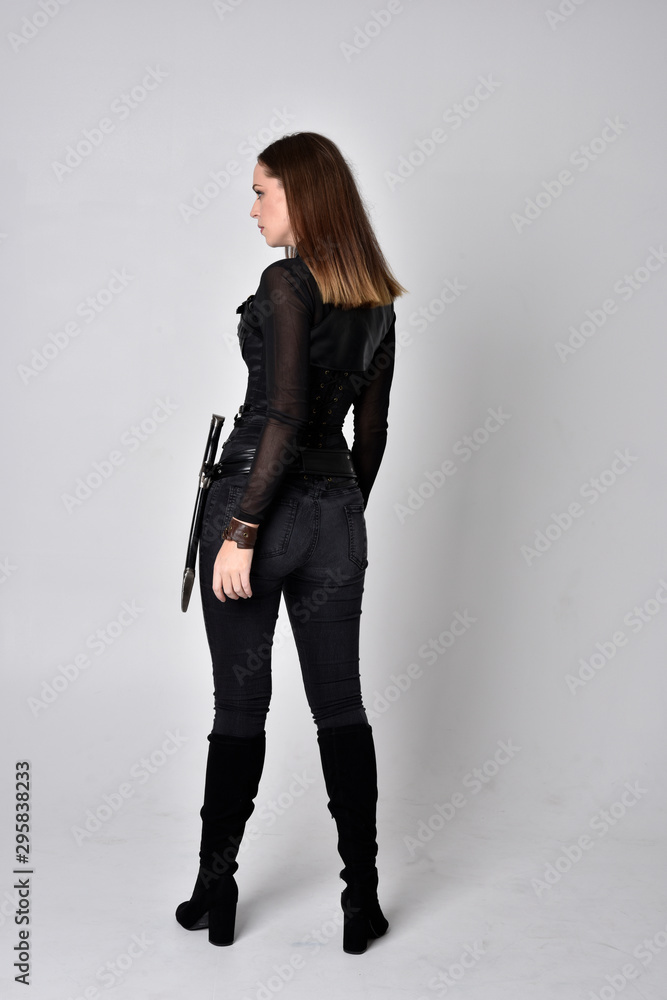 full length portrait of a pretty brunette woman wearing black leather fantasy costume with a dagger. standing pose, facing away from camera on a studio background. full length portrait of a pretty brunette woman wearing black leather fantasy costume with a dagger. standing pose, facing away from camera on a studio background.
