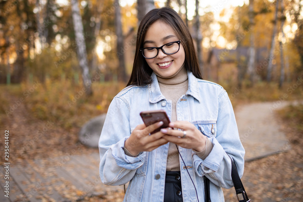 Young girl smiles and prints a message in a smartphone on a background of autumn nature Young girl smiles and prints a message in a smartphone on a background of autumn nature
