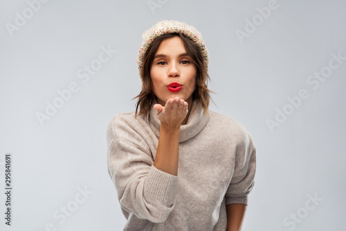 christmas, season and people concept - happy young woman in knitted winter hat and sweater sending air kiss over grey background