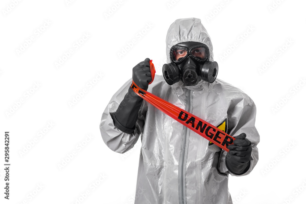 A man in a gray suit with a hood on his head with a yellow radiation sign, with a protective gas mask, posing while standing on a white background, winding a red protective tape around his hand. A man in a gray suit with a hood on his head with a yellow radiation sign, with a protective gas mask, posing while standing on a white background, winding a red protective tape around his hand.