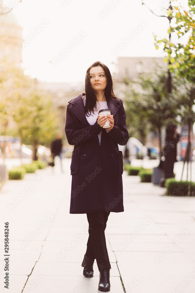 Beautiful serious smart brunette girl holding cup of coffee in hands goes walking down street of St. Petersburg in city center. Charming thoughtful woman with long dark hair wanders alone Beautiful serious smart brunette girl holding cup of coffee in hands goes walking down street of St. Petersburg in city center. Charming thoughtful woman with long dark hair wanders alone