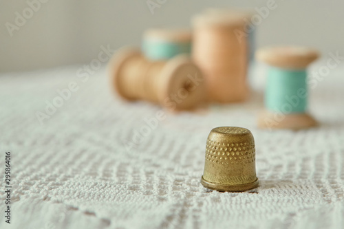 Vintage brass thimble close-up.  It is on an old handmade knitted tablecloth.  In the background vintage wooden bobbins with sewing threads photo