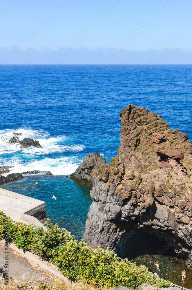 Tourists swimming in natural pools in the Atlantic ocean in Seixal, Madeira island, Portugal. Pool surrounded by volcanic rocks from the open sea. View from above. Summer vacation destination