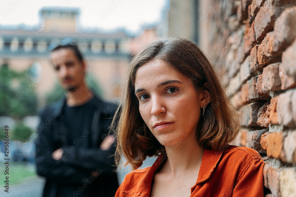 Φωτογραφία Girl looking at camera with an assertive look and a brick wall on the side and with a boy and a building out of focus in the background