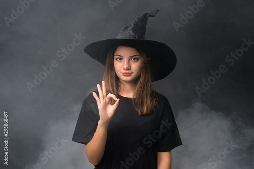 Girl with witch costume for halloween parties over isolated dark background showing an ok sign with fingers