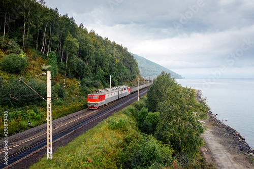 Freight train on the railroad of Trans-Siberian Railway on the shore of Baikal Lake with green forest trees and cloudy sky. East Siberian Railway in Buryatia, Siberia, Russia