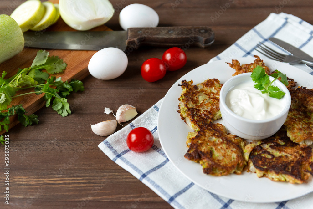 plate with zucchini pancakes with sour cream and herbs on a wooden background, knife, fork, fresh vegetables, cherry tomatoes, garlic plate with zucchini pancakes with sour cream and herbs on a wooden background, knife, fork, fresh vegetables, cherry tomatoes, garlic
