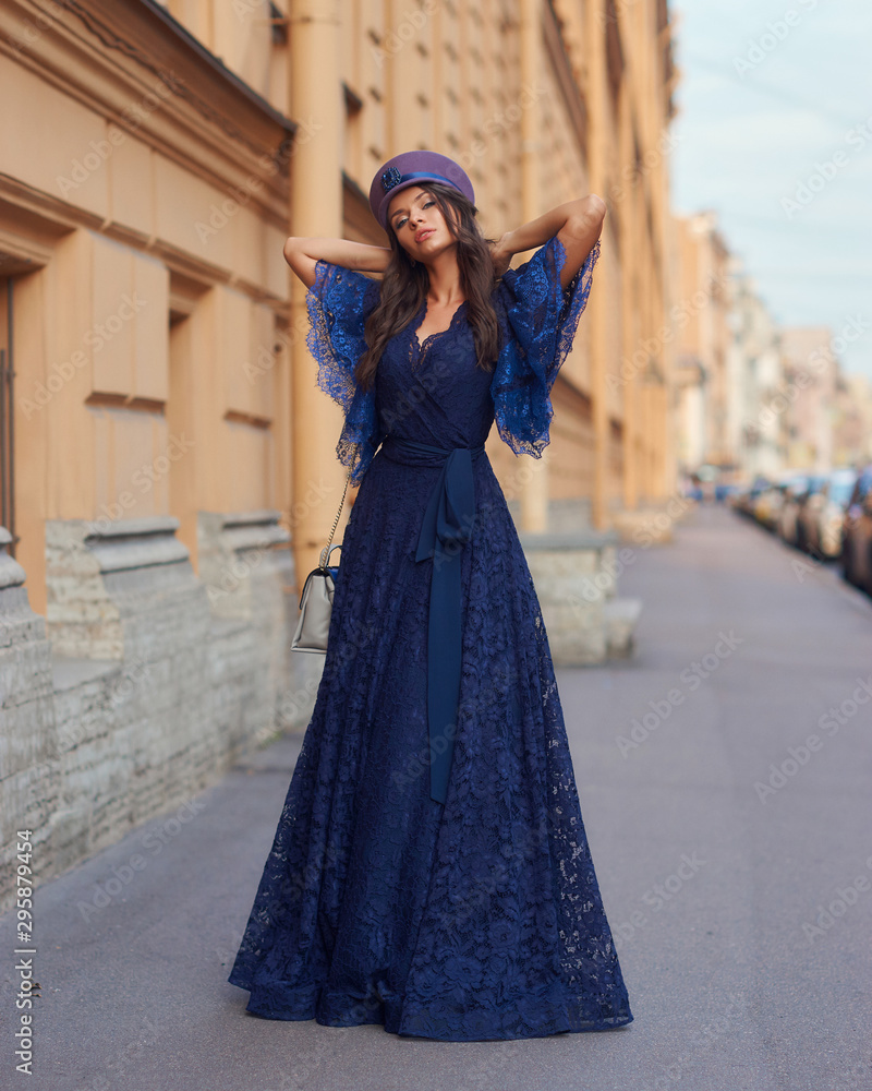 Full length outdoor portrait of young beautiful elegant woman in long blue evening dress and purple hat standing and posing at old city street on a sunny evening day