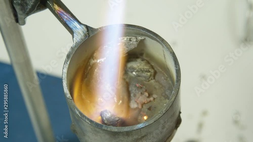 Melting silvery metal in a metal tin with a hot flame. Burner flame melting pieces of left over metal in small crucible / melting pot.