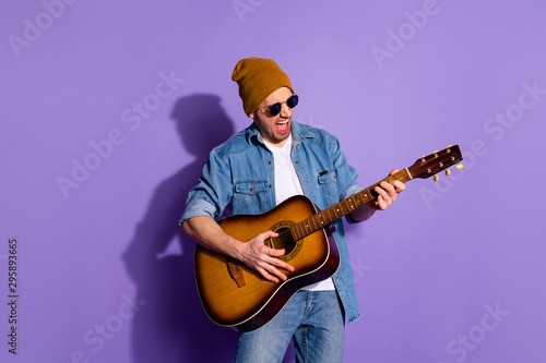 Photo of cheerful rude attractive handsome man wearing cap holding guitar with hands playing musician instrument wearing spectacles isolated over purple vivid color background
