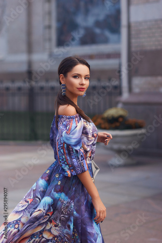 Outdoor portrait of stylish woman wearing colorful blue dress. Brunette caucasian woman with long hair in a tail walking down city street