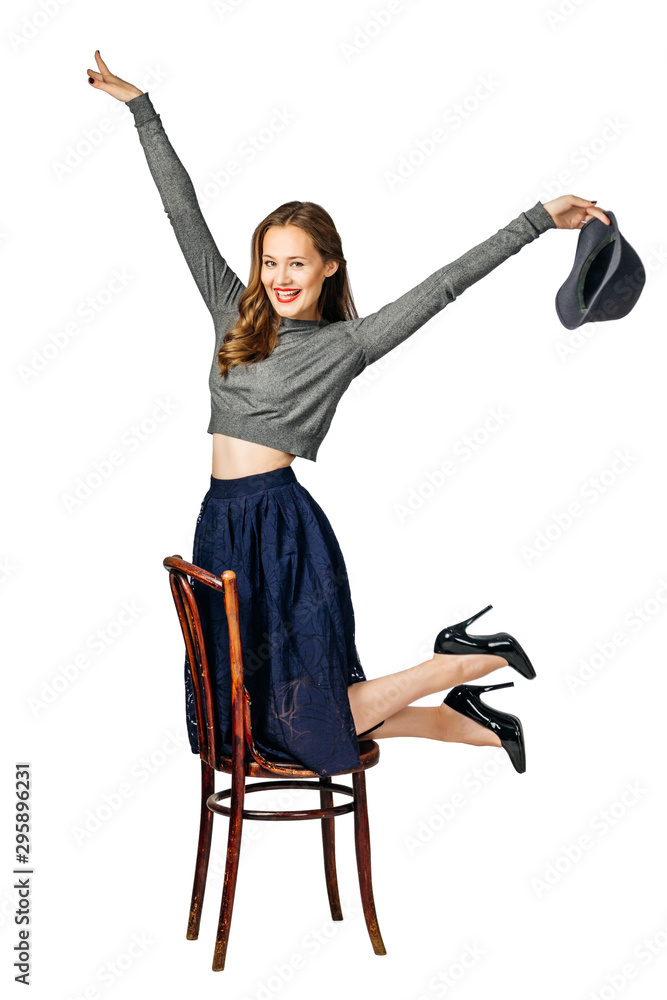 young beautiful cheerful girl in blue skirt and gray jumper kneels on chair and holds gray hat in hand and laughs. isolated on white background young beautiful cheerful girl in blue skirt and gray jumper kneels on chair and holds gray hat in hand and laughs. isolated on white background