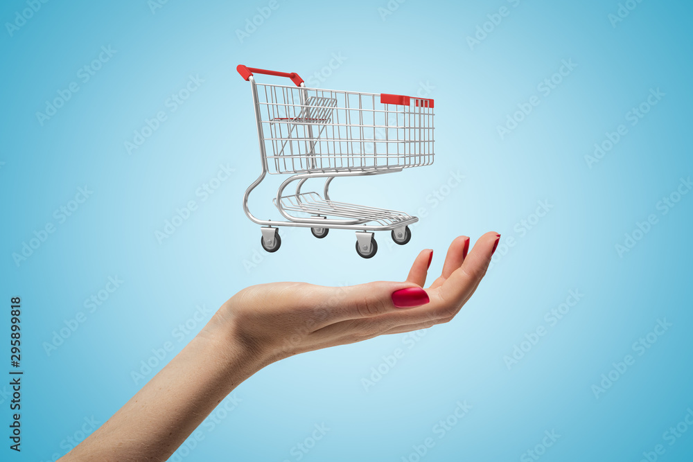 Side closeup of woman's hand facing up and levitating small supermarket trolley on light blue gradient background.