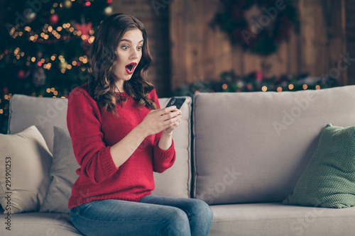 Photo of excited lady holding telephone reading unexpected best friends congratulation letters sitting couch in decorated garland lights room indoors wearing red sweater