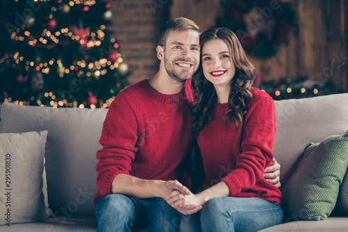 Happy new year. Photo of emotional couple meeting christmas in decorated garland lights room sitting close on cosy sofa indoors wearing red pullovers