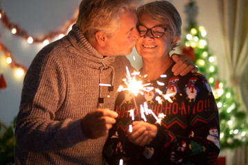 An affectionate kiss between elderly wife and husband who celebrate Christmas with sparks. Lights and Christmas tree in the background