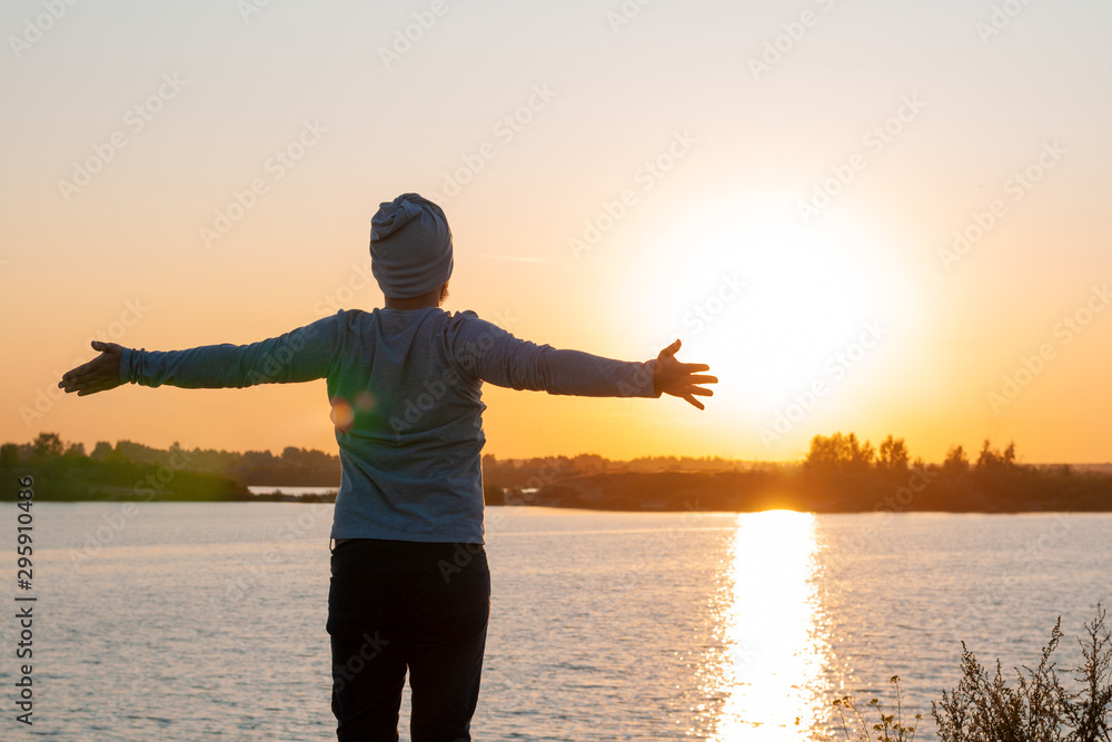 A young man, a hipster, stands on a lake at sunset, enjoys the view while traveling, desolation, enjoyment, harmony.