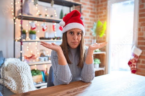 Young beautiful woman wearing christmas hat sitting at the table at home clueless and confused expression with arms and hands raised. Doubt concept.