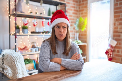 Young beautiful woman wearing christmas hat sitting at the table at home skeptic and nervous, disapproving expression on face with crossed arms. Negative person.