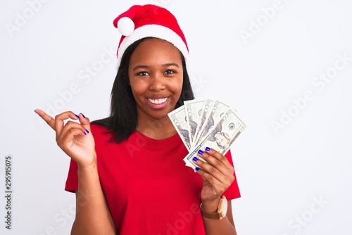 Young african american woman wearing christmas hat and holding bunch of dollars banknotes very happy pointing with hand and finger to the side © Krakenimages.com