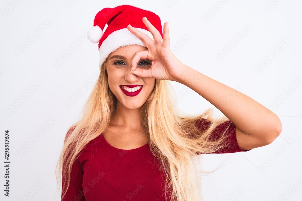 Young beautiful woman wearing Christmas Santa hat over isolated white background doing ok gesture with hand smiling, eye looking through fingers with happy face. Young beautiful woman wearing Christmas Santa hat over isolated white background doing ok gesture with hand smiling, eye looking through fingers with happy face.