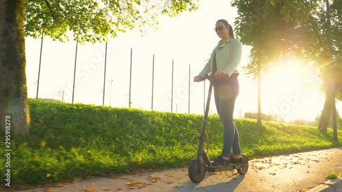 SLOW MOTION, LENS FLARE, LOW ANGLE: Girl having fun riding an electric scooter on a sunny evening. Cinematic shot of golden sunshine illuminating the fall colored avenue as woman rides an e-scooter