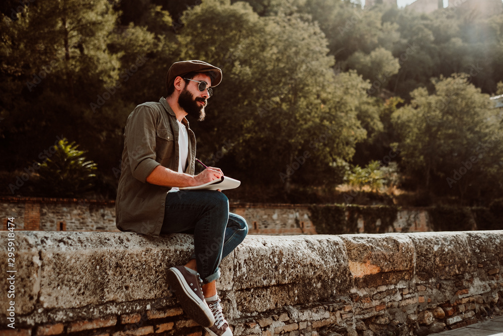 Fototapeta premium .Young man sitting in the street of Granada, Spain, drawing in his travel notebook. Enjoying the sunset, developing his creativity. Relaxed and carefree. Lifestyle.