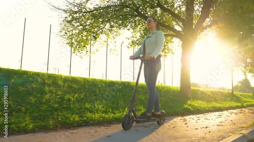 LOW ANGLE, SUN FLARE: Young woman in jeans rides a high tech e-scooter along an avenue in the autumn. Caucasian girl having fun riding an electric scooter down empty sidewalk on a sunny fall evening.