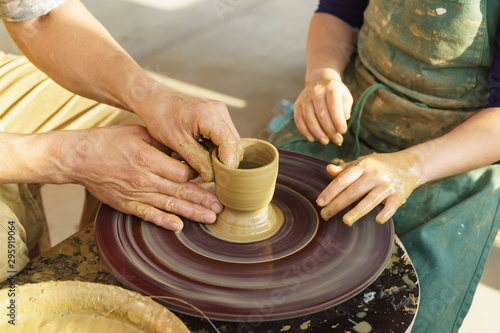 The hands of an adult man show the child how to make a jug from a piece of clay on a Potters wheel