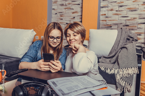 Portrait of beautiful smiling woman and her teenage daughter in jeans shirt using phone and watching funny video