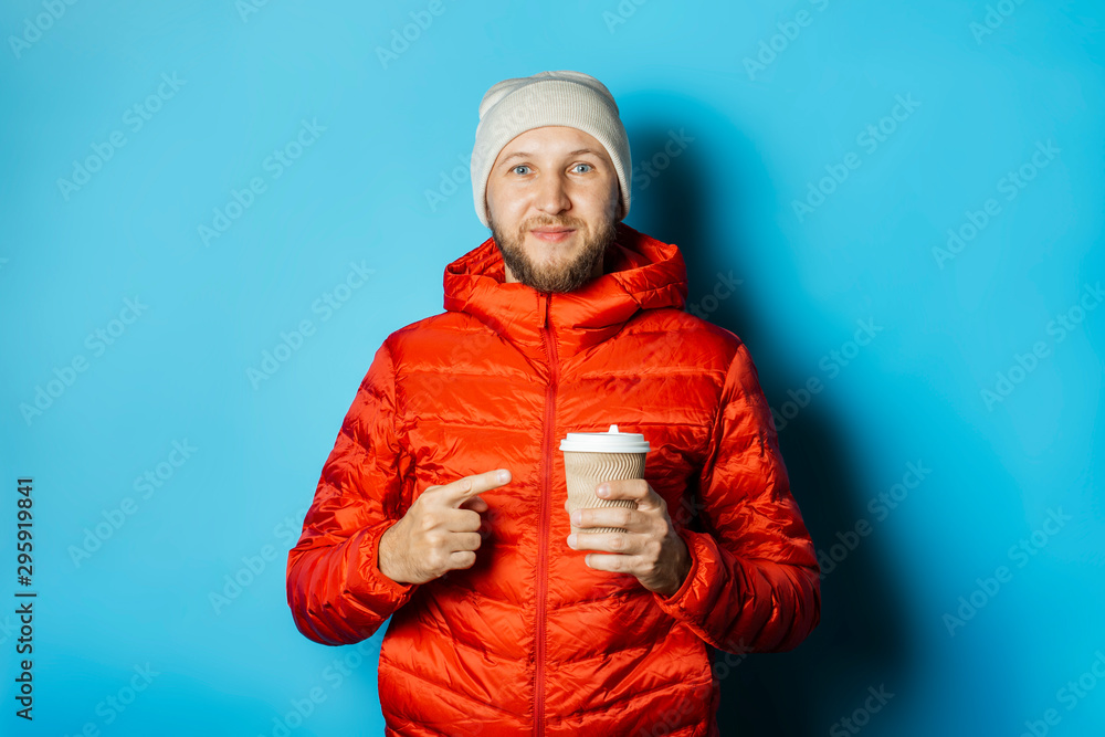 Man in a hat and a red jacket holds a cup of hot coffee on a blue background. Warming drinks, winter, autumn Man in a hat and a red jacket holds a cup of hot coffee on a blue background. Warming drinks, winter, autumn