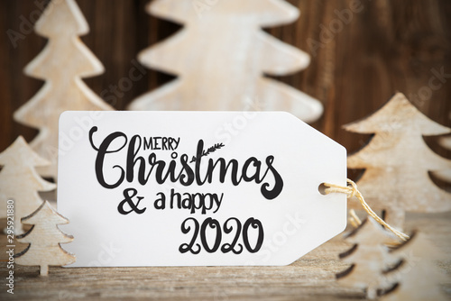 Label With English Text Merry Christmas And Happy 2020. White Wooden Christmas Tree As Decoration. Brown Wooden Background