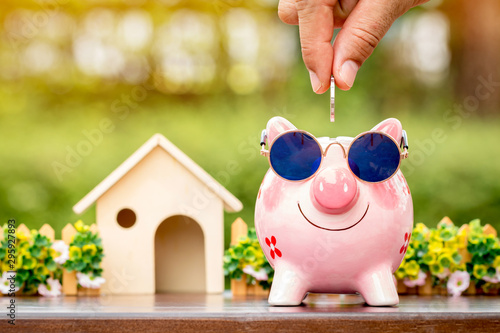 Businessman hand hold a coin is save to the piggy bank with growing a interest and wooden home model for family in the public park, Business investment and loan for real estate in the future concept. photo