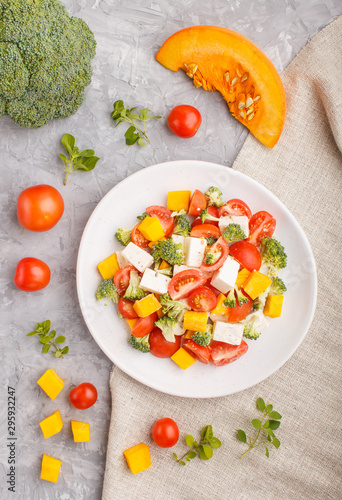 Vegetarian salad with broccoli, tomatoes, feta cheese, and pumpkin on white ceramic plate on a gray concrete background, top view.