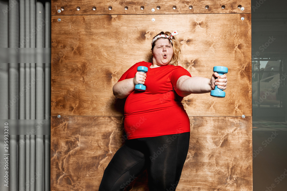 Funny fat girl dressed in the sportswear and with a bandage on her head  stands with dumbbells on the background of a wooden wall in the gym Stock  Photo | Adobe Stock