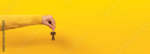 female hand holding house keys, suggesting, over yellow background, panoramic image.