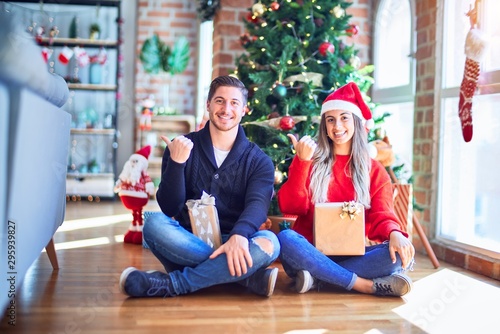 Young couple wearing santa claus hat sitting on the floor around christmas tree at home smiling with happy face looking and pointing to the side with thumb up.