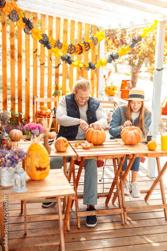 Halloween Preparaton Concept. Young couple sitting at table outdoors making jack-o'-lantern carving pumpkins joyful