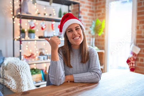 Young beautiful woman wearing christmas hat sitting at the table at home smiling with happy face looking and pointing to the side with thumb up.