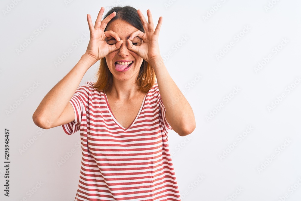 Beautiful redhead woman wearing casual striped red t-shirt over isolated background doing ok gesture like binoculars sticking tongue out, eyes looking through fingers. Crazy expression.