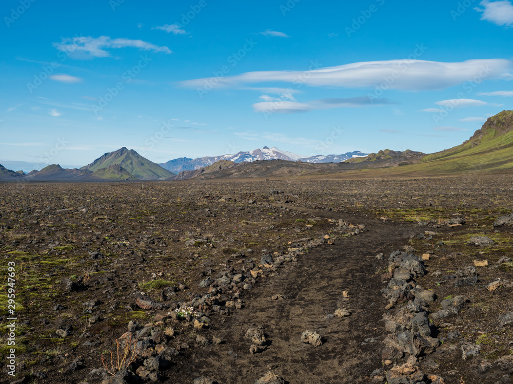 Icelandic lava desert landscape with footpath of Laugavegur hiking trail with view on Tindfjallajokull glacier mountains and green hills. Fjallabak Nature Reserve, Iceland. Summer blue sky