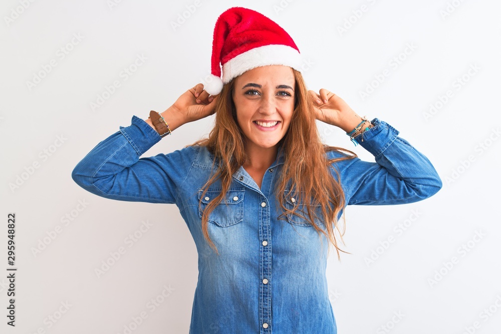 Young beautiful redhead woman wearing christmas hat over isolated background Smiling pulling ears with fingers, funny gesture. Audition problem Young beautiful redhead woman wearing christmas hat over isolated background Smiling pulling ears with fingers, funny gesture. Audition problem