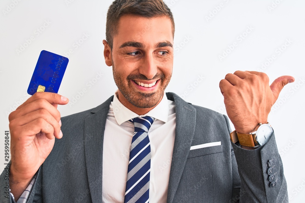 Young handsome business man holding credit card over isolated background pointing and showing with thumb up to the side with happy face smiling Young handsome business man holding credit card over isolated background pointing and showing with thumb up to the side with happy face smiling