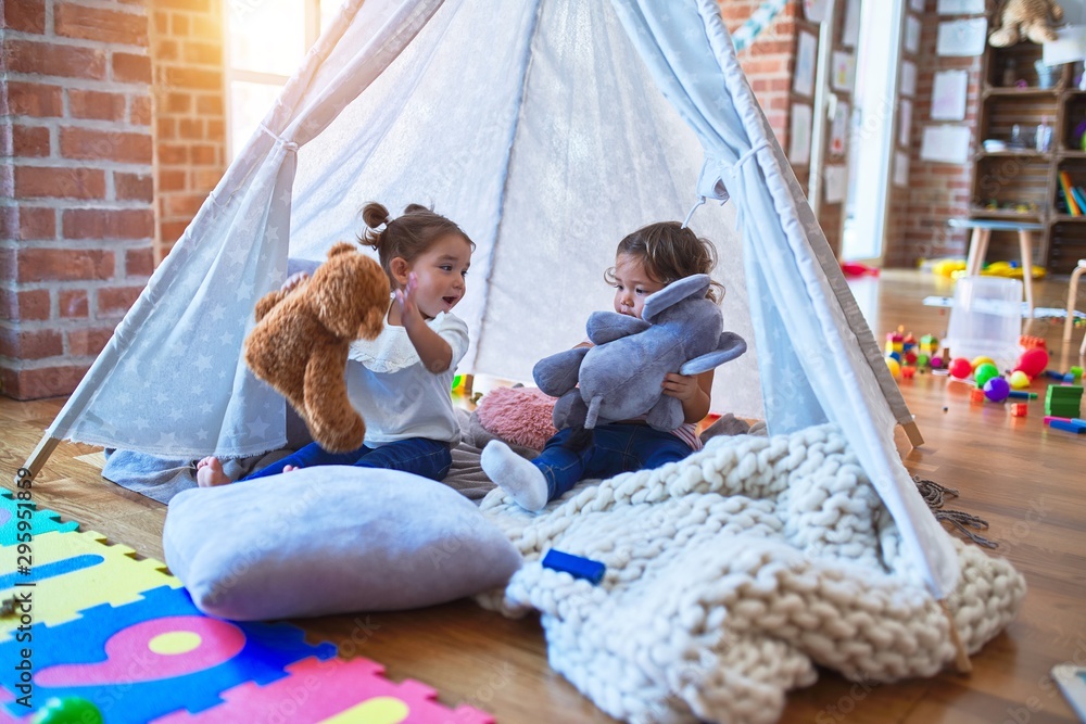 Adorable toddlers lying down over blanket inside tipi smiling and playing with doll at kindergarten