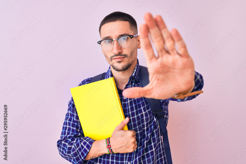 Young handsome student man holding a book over isolated background with open hand doing stop sign with serious and confident expression, defense gesture Young handsome student man holding a book over isolated background with open hand doing stop sign with serious and confident expression, defense gesture