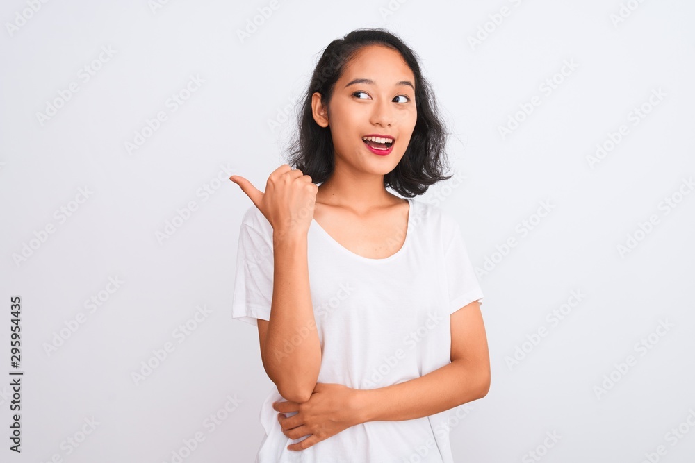 Young chinese woman wearing casual t-shirt standing over isolated white background smiling with happy face looking and pointing to the side with thumb up. Young chinese woman wearing casual t-shirt standing over isolated white background smiling with happy face looking and pointing to the side with thumb up.